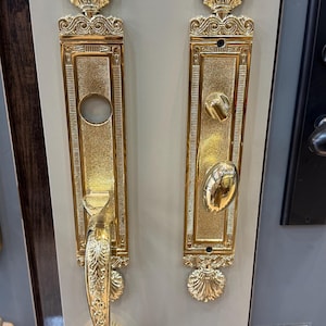 Puede incluir: Dos manijas doradas ornamentadas con un diseño de concha. Las manijas tienen forma rectangular con detalles intrincados y un acabado pulido. Una manija tiene una palanca y la otra un pomo. Adecuado para puertas.