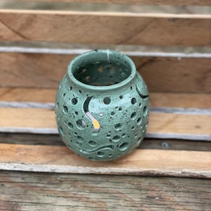 May include: A spherical ceramic candle holder in a light green glaze with dark speckles. The surface features numerous circular cutouts of varying sizes and curved, wave-like slits that allow light to pass through the hollow interior structure.