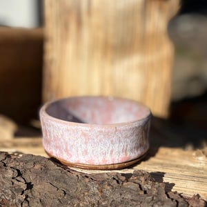 May include: A small, round ceramic bowl featuring a mottled glaze with shades of soft pink and white. The bowl has a smooth, slightly curved rim and a distinct, unglazed brown base that contrasts with the lighter, textured finish of the upper body.