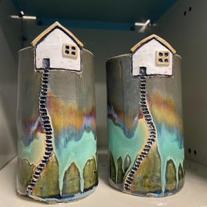 May include: Two ceramic vases with a house design. Each vase is cylindrical with a white house on top and a black ladder leading down. The vases have a color gradient of green, blue, and brown.