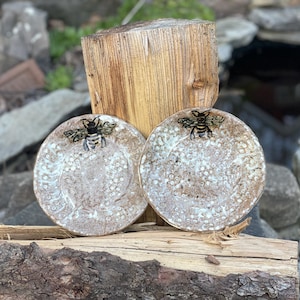May include: Two handmade ceramic plates with a bee design. The plates are round, with a textured surface and a brown and white color scheme. The bee is black and gold. The plates are displayed on a wooden surface.