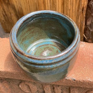 May include: A small, round ceramic cup with a glossy glaze featuring a blend of deep blue, teal, and earthy brown tones. The interior shows visible circular throwing lines and a smooth, reflective surface, while the exterior has a thick, rounded rim.