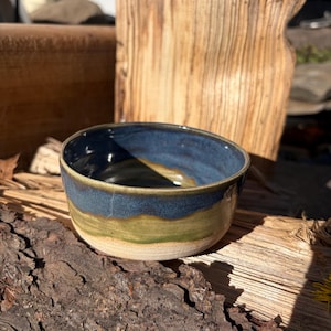 May include: A handmade ceramic bowl with a blue, green, and cream color scheme. The bowl has a rounded shape with a dark blue rim, a green midsection, and a cream-colored base. The bowl is sitting on a piece of wood.