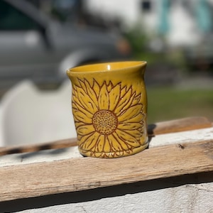 May include: A yellow ceramic mug featuring a prominent, embossed sunflower design on its exterior. The sunflower has detailed brown-toned petals and a textured center, contrasting against the smooth, glossy yellow glaze of the cylindrical vessel.