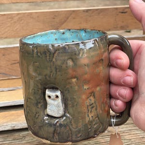 May include: A handmade ceramic mug with a textured, mottled brown and orange exterior glaze. The interior is a bright turquoise blue. A small, sculpted white owl figure is attached to the side of the mug, appearing to peek out from a hollow.