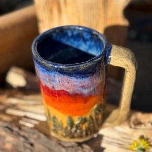 May include: A handmade ceramic mug with a landscape design. The mug features a blue rim, transitioning to red and orange hues, with a forest scene at the base. The handle is a tan color.