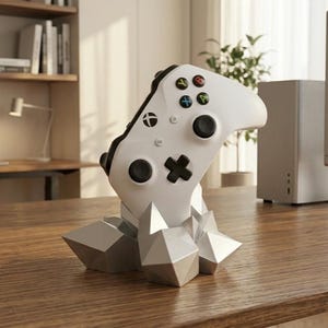 Peut inclure: Une manette Xbox blanche repose sur un support géométrique gris. La manette comporte des boutons noirs et des boutons d'action colorés. Le support a un design à facettes, ressemblant à un amas de cristaux. L'ensemble est sur un bureau en bois.