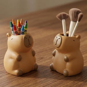 Puede incluir: Dos organizadores de escritorio con forma de carpincho de color beige. Uno contiene crayones de colores, el otro brochas de maquillaje. Los organizadores tienen un acabado liso, con detalles marrones para los ojos, la nariz y los pies. Están sobre una superficie de madera.