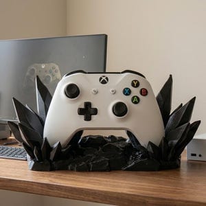 Può includere: Un controller Xbox bianco poggia su un supporto nero a forma di cristallo. Il controller ha pulsanti colorati e un pad direzionale nero. Il supporto è su una superficie di legno, con un monitor di computer e una tastiera sullo sfondo.