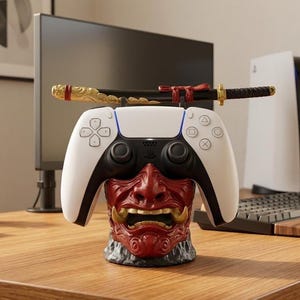 Peut inclure: Une manette PlayStation 5 blanche et noire repose sur un support en forme de masque Oni rouge et doré. Un katana décoratif repose sur le dessus de la manette. La manette et le support sont sur un bureau en bois, avec un écran d'ordinateur et une console PS5 en arrière-plan.