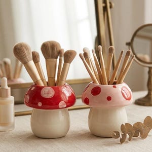 Op de afbeelding: Twee paddenstoelvormige keramische borstelhouders, één rood met witte stippen, de andere roze met rode stippen, elk gevuld met make-up borstels. De houders staan op een lichtgekleurd oppervlak, met een spiegel en andere schoonheidsproducten op de achtergrond.