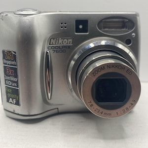 May include: Silver Nikon COOLPIX 7600 digital camera with a zoom lens. The camera has a 7.1-megapixel resolution and a 3x zoom. The lens has a focal length of 7.8-23.4mm and an aperture of f/2.8-4.9.