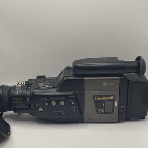 Peut inclure: Caméscope Kyocera Finemovie 8 noir et gris. L'appareil photo est équipé d'un objectif zoom, de divers boutons et d'une poignée. L'appareil photo est posé sur une surface blanche.