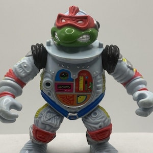 May include: A Teenage Mutant Ninja Turtle action figure in a gray and blue spacesuit. The figure has a green head with a red mask and a detailed chest plate with the word "OOZE". The figure is a collectible toy.