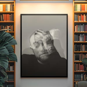 Op de afbeelding: Zwart-wit portretprint van een man met zijn hand op zijn hoofd. De afbeelding is zwart omlijst en hangt boven een bank. De achtergrond omvat boekenplanken en een grote potplant.