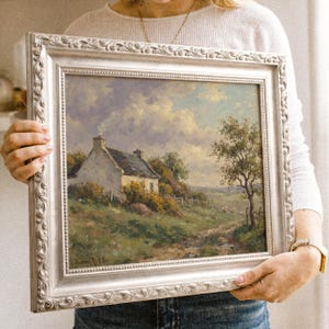 Vintage oil painting of a white stone cottage with a gray roof on a green hillside, surrounded by flowering bushes, a lone tree, and a dirt path under a cloudy sky, displayed in an ornate gold frame.