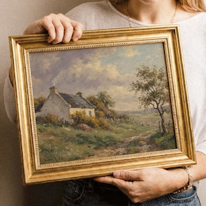 Vintage oil painting of a white stone cottage with a gray roof on a green hillside, surrounded by flowering bushes, a lone tree, and a dirt path under a cloudy sky, displayed in an ornate gold frame.