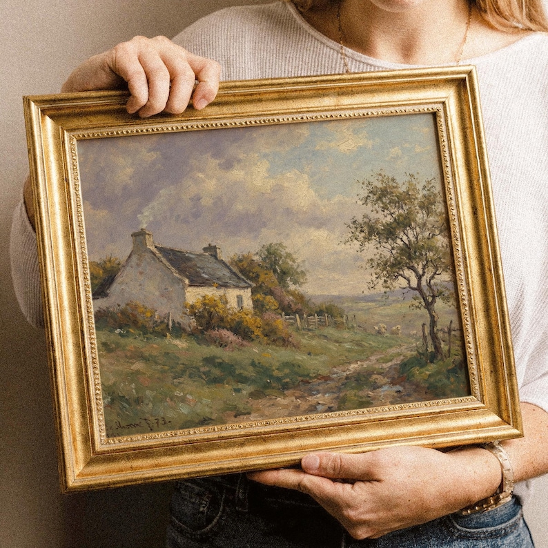 Vintage oil painting of a white stone cottage with a gray roof on a green hillside, surrounded by flowering bushes, a lone tree, and a dirt path under a cloudy sky, displayed in an ornate gold frame.