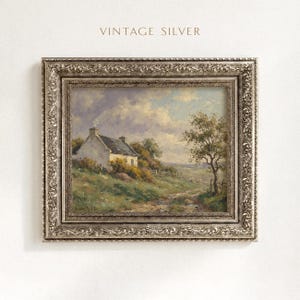 Vintage oil painting of a white stone cottage with a gray roof on a green hillside, surrounded by flowering bushes, a lone tree, and a dirt path under a cloudy sky, displayed in an ornate gold frame.