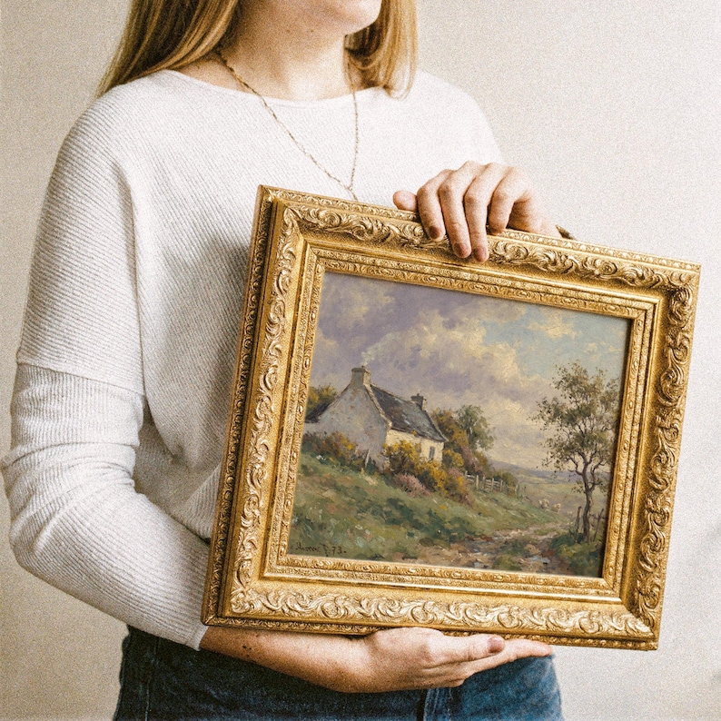 Vintage oil painting of a white stone cottage with a gray roof on a green hillside, surrounded by flowering bushes, a lone tree, and a dirt path under a cloudy sky, displayed in an ornate gold frame.