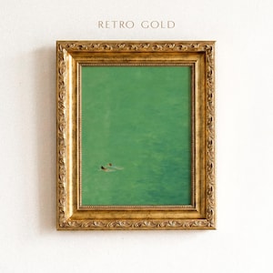 Op de afbeelding: Een rechthoekige wandkunstprint in een decoratieve, sierlijke gouden lijst met de tekst RETRO GOLD erboven. Het kunstwerk heeft een effen, getextureerde groene achtergrond met twee kleine figuren die in het linksonder gedeelte zwemmen.