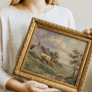 Vintage oil painting of a white stone cottage with a gray roof on a green hillside, surrounded by flowering bushes, a lone tree, and a dirt path under a cloudy sky, displayed in an ornate gold frame.