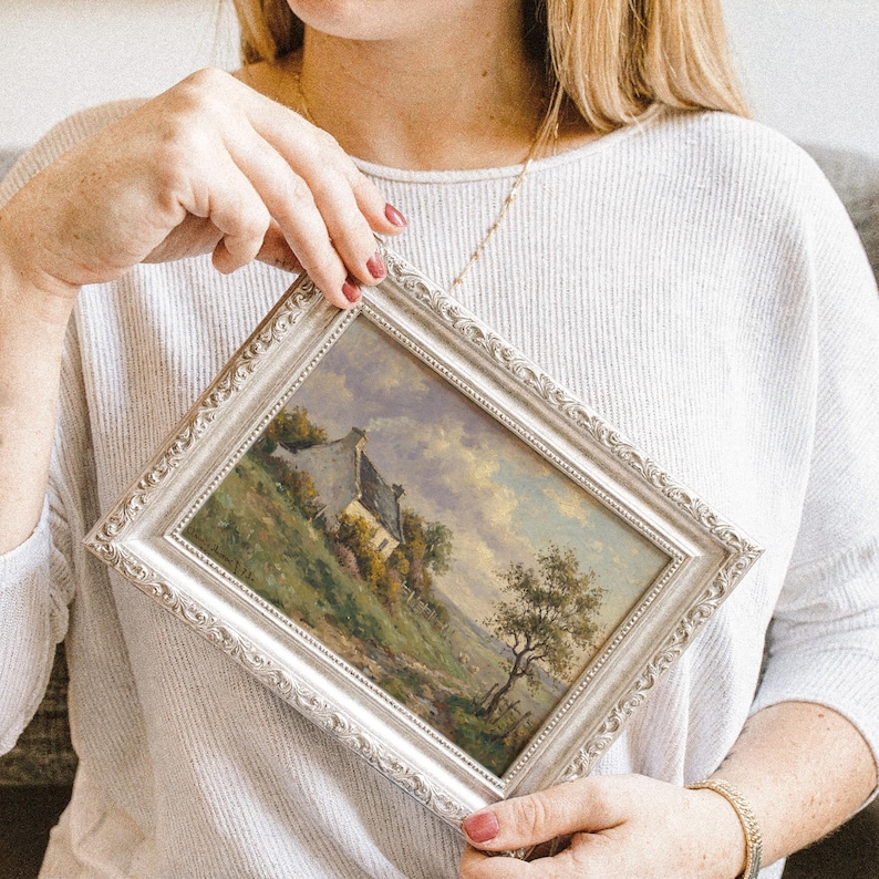 Vintage oil painting of a white stone cottage with a gray roof on a green hillside, surrounded by flowering bushes, a lone tree, and a dirt path under a cloudy sky, displayed in an ornate gold frame.