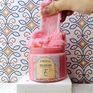 May include: A clear plastic jar filled with pink textured slime topped with small pink sprinkles. The label features the text Cherry Blossom and Y & M Slimes with a floral design. A portion of the pink slime is being pulled upward from the jar.