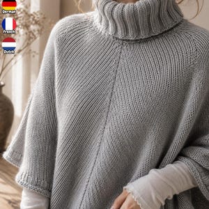 Puede incluir: Un poncho de punto gris claro con un cuello alto de canalé prominente y un patrón de puntadas en diagonal en la parte delantera. La prenda tiene mangas cortas y anchas con puños de canalé y se lleva sobre una camiseta blanca de manga larga.
