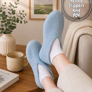 May include: Light blue felted slippers with a soft, textured finish, worn with white socks. The image includes a coffee mug, a vase with greenery, and a book on a wooden table. Text on the image says "Cozy Felted Slippers Knit PDF".
