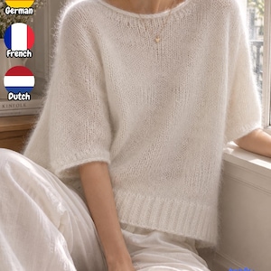 May include: A white, short-sleeved, loose-fitting jumper with a wide, rounded neckline and a ribbed hem. The garment features a soft, fuzzy, semi-sheer knit texture. A delicate gold chain necklace with a small heart pendant is worn around the neck.