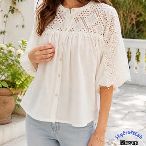 Puede incluir: Una blusa blanca con botones, un canesú de encaje detallado y ribetes de encaje a juego en las mangas tres cuartos. La prenda tiene un cuello redondo y un corte holgado. El texto SkyCraftLab Elowen Lace Yoke Blouse es visible en la imagen.
