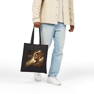 Op de afbeelding: Zwarte draagtas met een artistiek portret van een vrouw met vloeiend haar en de tekst "Carrying the weight of those before me". Het kunstwerk is in goud-, bruin- en crèmetinten.