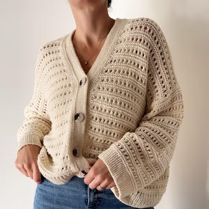 以下が含まれることがあります： クリーム色のオープンニットカーディガン、Vネック、ボタン留め。長袖セーターはブルージーンズと組み合わせています。カーディガンはテクスチャードニットのデザインで、多目的に使えるレイヤリングアイテムです。