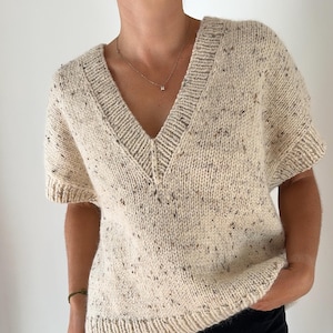 May include: A cream-colored knitted sweater with a speckled pattern and a v-neck. The sweater has short sleeves and a ribbed hem.