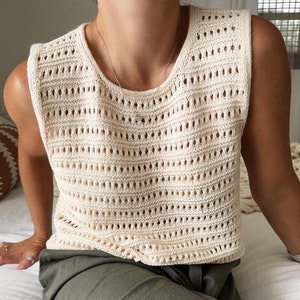 May include: A cream-colored crochet tank top with a loose fit and a delicate open-weave pattern. The top is sleeveless and has a round neckline.
