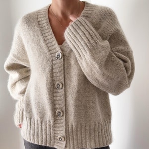 May include: A beige, chunky-knit cardigan with a V-neck and button closure. The buttons have a decorative design. The sleeves are long and the cardigan appears to be made of a soft, warm material. The cardigan is worn over a white top.