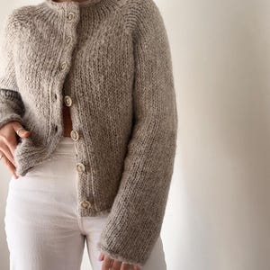 Puede incluir: Una rebeca de punto grueso en tono beige jaspeado con cuello redondo, mangas largas y cierre frontal con seis botones circulares de color claro. La prenda se lleva sobre un pantalón blanco, mostrando un hilo con textura similar a la lana.