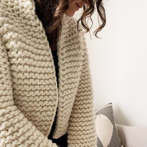 K&ouml;nnte beinhalten: Nahaufnahme eines cremefarbenen Strickjackens mit lockerer, grober Strickart. Der Cardigan ist kurz und hat einen V-Ausschnitt.