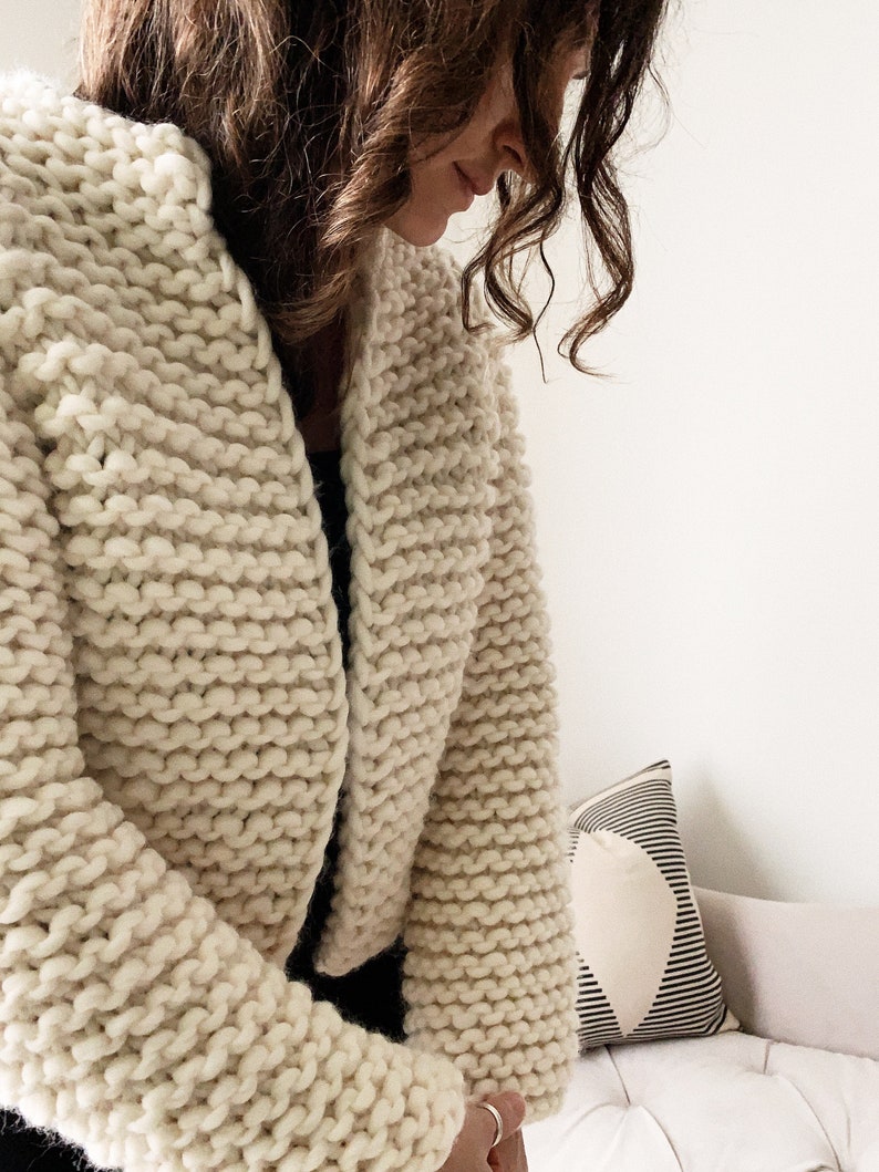 K&ouml;nnte beinhalten: Nahaufnahme eines cremefarbenen Strickjackens mit lockerer, grober Strickart. Der Cardigan ist kurz und hat einen V-Ausschnitt.