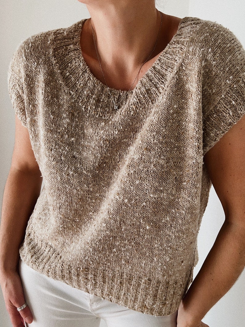 May include: A beige knitted sleeveless top with a speckled texture. The top has a round neckline and a slightly cropped hem.