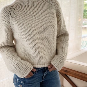 May include: A cream-colored chunky knit sweater with a mock neck and long sleeves. The sweater is made with a thick yarn and has a relaxed fit.