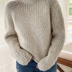 May include: A cream-colored chunky knit sweater with a high neck and long sleeves.