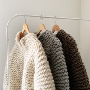 K&ouml;nnte beinhalten: Drei Strickpullover h&auml;ngen an einem wei&szlig;en Gestell. Die Pullover sind in den Farben Wei&szlig;, Grau und Braun.
