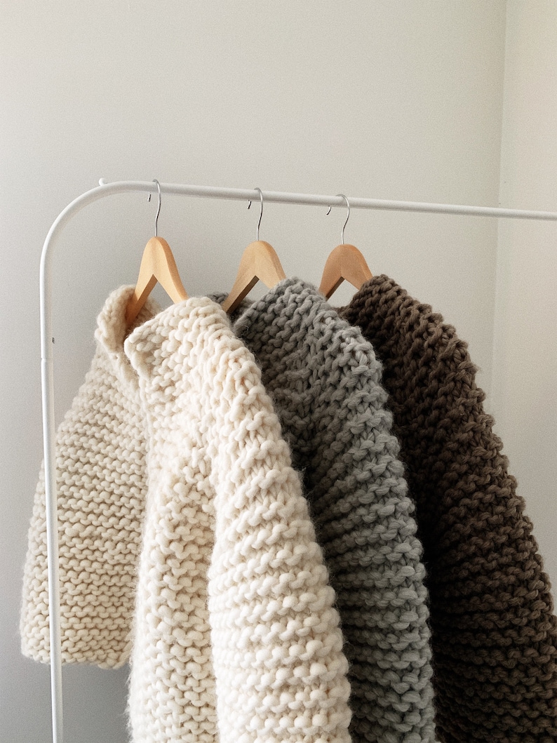 K&ouml;nnte beinhalten: Drei Strickpullover h&auml;ngen an einem wei&szlig;en Gestell. Die Pullover sind in den Farben Wei&szlig;, Grau und Braun.