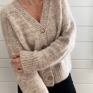 May include: A beige cardigan with a v-neck and button closure. The cardigan is made of a soft, fuzzy material and has a speckled pattern.