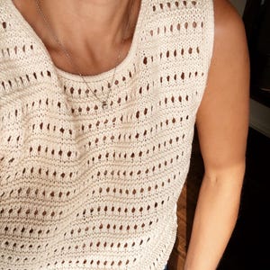 May include: A white crochet tank top with a delicate open-stitch pattern. The top is sleeveless and has a scoop neck.