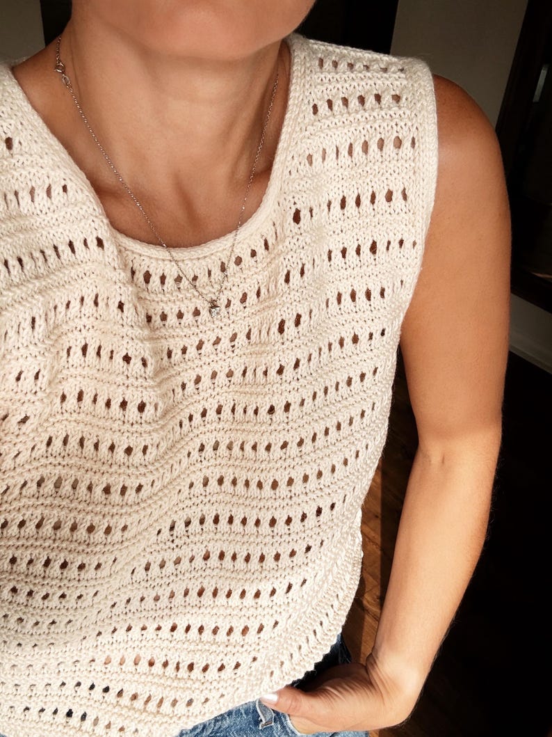 May include: A white crochet tank top with a delicate open-stitch pattern. The top is sleeveless and has a scoop neck.