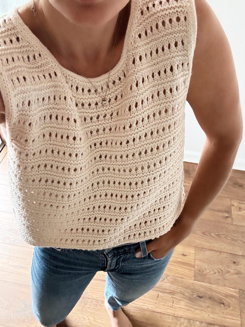 May include: A crocheted sleeveless top in a cream color with a delicate open-stitch pattern. The top is cropped and has a relaxed fit.