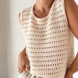 May include: A cream-colored crochet tank top with a loose fit and a round neckline. The top has a delicate open-weave pattern.